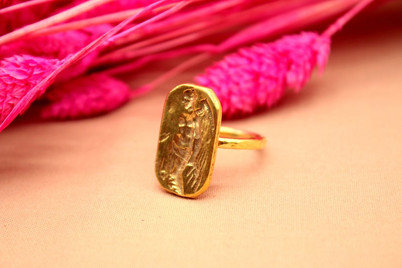 May include: A gold ring with a rectangular design featuring an embossed image of a winged figure. The ring is set against a pink background.