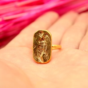 May include: A gold ring with a rectangular design featuring a raised image of an angel with wings.
