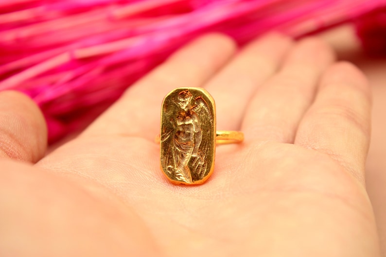 May include: A gold ring with a rectangular design featuring a raised image of an angel with wings.