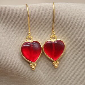 May include: A pair of gold heart-shaped earrings with red glass inserts. The earrings have a dangling design with small gold beads at the bottom.