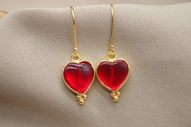 May include: A pair of gold heart-shaped earrings with red glass inserts. The earrings have a dangling design with small gold beads at the bottom.