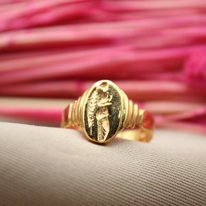 Op de afbeelding: Een gouden ring met een ovaalvormig ontwerp met een gedetailleerd figuur. De ring ligt op een roze achtergrond.