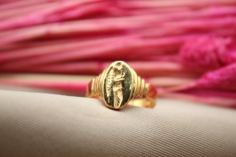 Op de afbeelding: Een gouden ring met een ovaalvormig ontwerp met een gedetailleerd figuur. De ring ligt op een roze achtergrond.