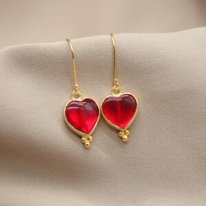 May include: A pair of gold heart-shaped earrings with red glass insets. The earrings have a delicate, dangling design.