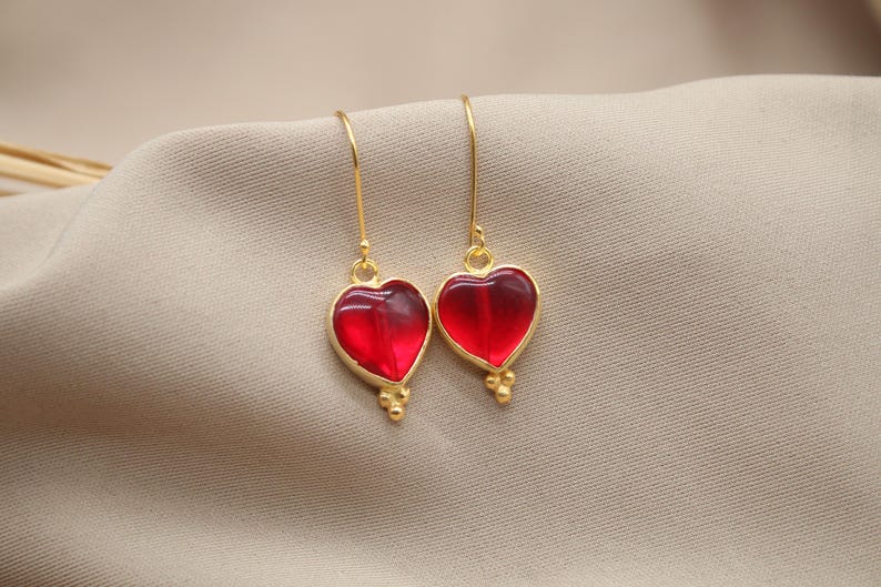 May include: A pair of gold heart-shaped earrings with red glass insets. The earrings have a delicate, dangling design.