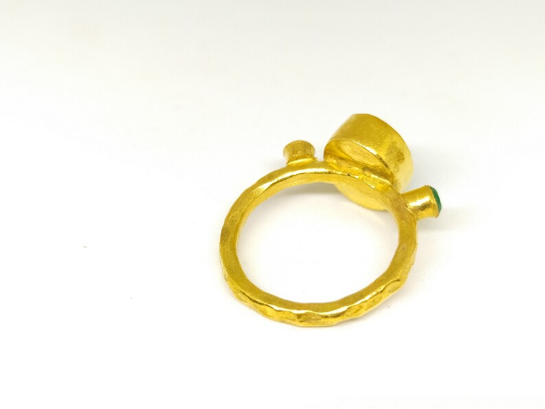 May include: A gold ring with a textured band and a large round gemstone. The ring has a smaller green gemstone on the side.