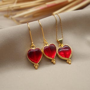 May include: A set of gold heart-shaped pendant earrings and necklace with red glass stones. The jewellery is on a light brown fabric background.