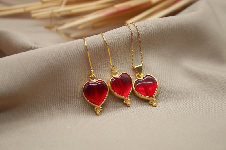 May include: A set of gold heart-shaped pendant earrings and necklace with red glass stones. The jewellery is on a light brown fabric background.