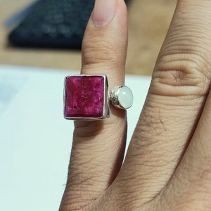 K&ouml;nnte beinhalten: Ein silberner Ring mit einem quadratischen, rosafarbenen Rubin-Edelstein und einem runden, wei&szlig;en Edelstein. Der Ring wird an einem Finger getragen.