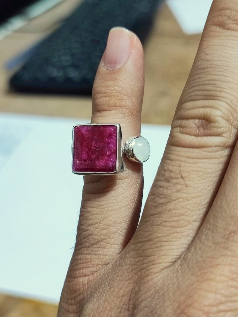 K&ouml;nnte beinhalten: Ein silberner Ring mit einem quadratischen, rosafarbenen Rubin-Edelstein und einem runden, wei&szlig;en Edelstein. Der Ring wird an einem Finger getragen.