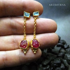 Op de afbeelding: Een paar gouden oorbellen met een hangende robijn en aquamarijn edelsteen. De oorbellen zijn gemaakt met een delicate gouden ketting en hebben een kleine, gefacetteerde aquamarijn aan de bovenkant en een grotere, gefacetteerde robijn aan de onderkant.