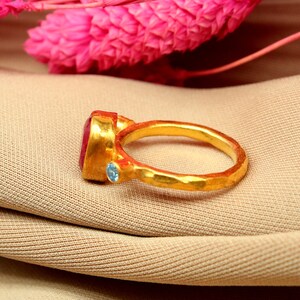 K&ouml;nnte beinhalten: Ein goldener Ring mit einem gro&szlig;en roten Edelstein und einem kleineren blauen Edelstein. Der Ring liegt auf einem braunen Stoffhintergrund.