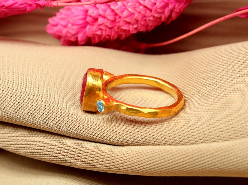 K&ouml;nnte beinhalten: Ein goldener Ring mit einem gro&szlig;en roten Edelstein und einem kleineren blauen Edelstein. Der Ring liegt auf einem braunen Stoffhintergrund.