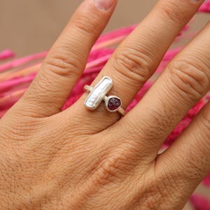 May include: A silver ring with a white pearl and a red gemstone. The ring is on a finger.
