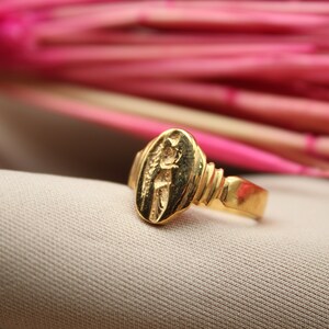 Op de afbeelding: Een gouden ring met een ovaalvormig ontwerp met een reli&euml;fbeeld van een persoon. De ring ligt op een lichtbruine stoffen achtergrond.