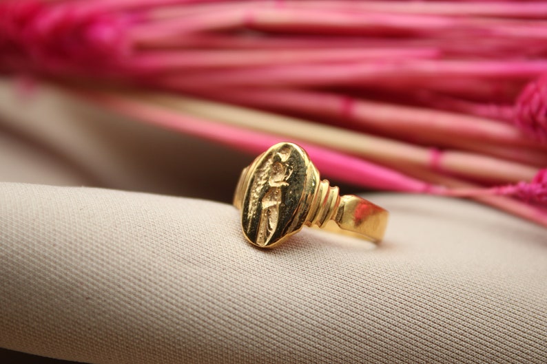 Op de afbeelding: Een gouden ring met een ovaalvormig ontwerp met een reli&euml;fbeeld van een persoon. De ring ligt op een lichtbruine stoffen achtergrond.