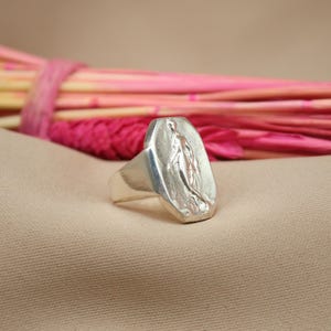 May include: A silver ring with a rectangular face featuring a detailed relief of a religious figure. The ring is set against a neutral beige backdrop with a blurred arrangement of pink and magenta decorative elements.