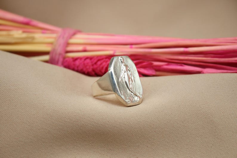 May include: A silver ring with a rectangular face featuring a detailed relief of a religious figure. The ring is set against a neutral beige backdrop with a blurred arrangement of pink and magenta decorative elements.