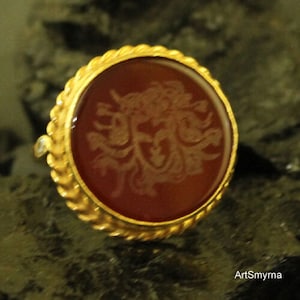 May include: A gold ring with a round, reddish-brown stone featuring an engraved design. The ring has a textured, rope-like border and a small, clear accent stone. The ring is set against a dark, textured background.
