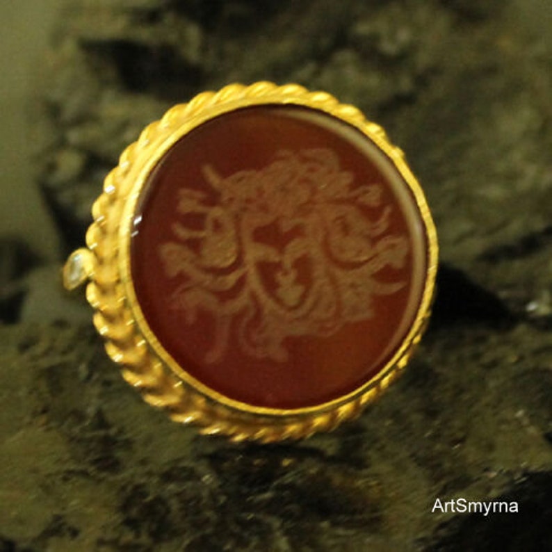 May include: A gold ring with a round, reddish-brown stone featuring an engraved design. The ring has a textured, rope-like border and a small, clear accent stone. The ring is set against a dark, textured background.