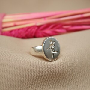 以下が含まれることがあります： 丸い顔にスタイリッシュな花のレリーフデザインが施されたシルバーリング。リングは薄い茶色の布地に置かれています。