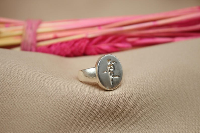 以下が含まれることがあります： 丸い顔にスタイリッシュな花のレリーフデザインが施されたシルバーリング。リングは薄い茶色の布地に置かれています。