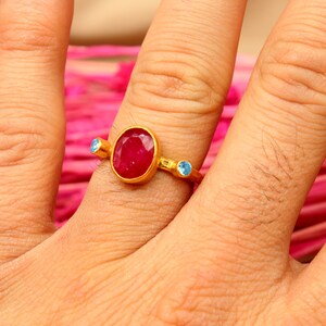 K&ouml;nnte beinhalten: Ein goldener Ring mit einem gro&szlig;en ovalen roten Edelstein und zwei kleineren blauen Edelsteinen an beiden Seiten.