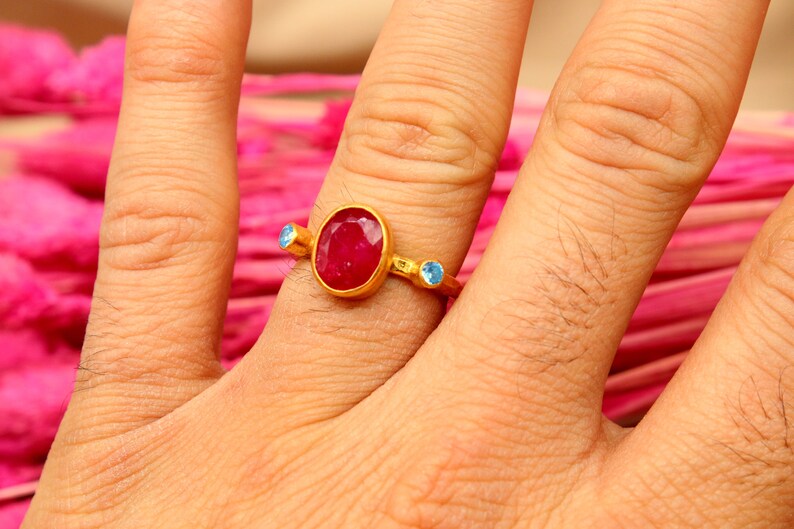K&ouml;nnte beinhalten: Ein goldener Ring mit einem gro&szlig;en ovalen roten Edelstein und zwei kleineren blauen Edelsteinen an beiden Seiten.