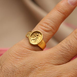 May include: A gold ring with a detailed design of a figure on the top. The ring is worn on a person's finger.