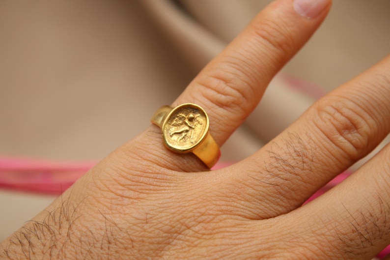 May include: A gold ring with a detailed design of a figure on the top. The ring is worn on a person's finger.