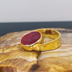Peut inclure: Une bague en or avec une pierre pr&eacute;cieuse rouge sertie au centre. La bague a une texture martel&eacute;e et un design simple.