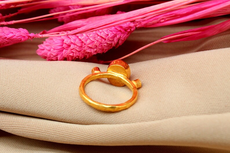 K&ouml;nnte beinhalten: Ein goldener Ring mit einem gro&szlig;en, runden, facettierten Edelstein. Der Ring hat eine strukturierte, geh&auml;mmerte Oberfl&auml;che und ein schlichtes, modernes Design.