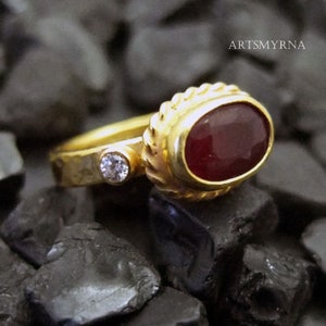 K&ouml;nnte beinhalten: Ein goldener Ring mit einem roten Edelstein und einem kleineren, klaren Edelstein. Der Ring liegt auf einem Bett aus schwarzen Steinen.