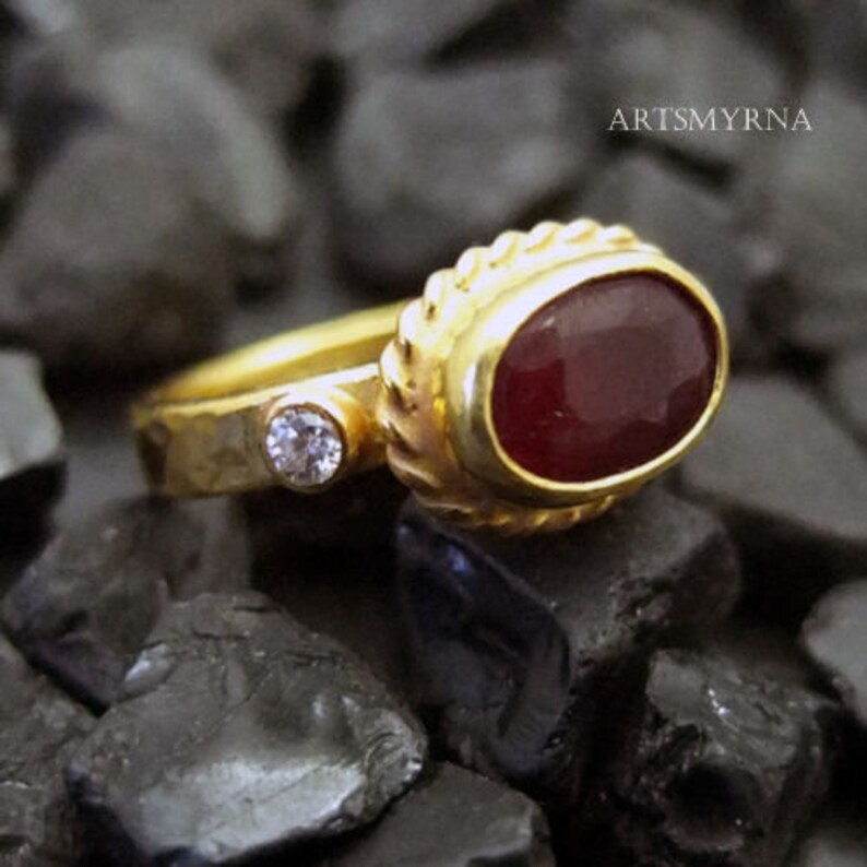 K&ouml;nnte beinhalten: Ein goldener Ring mit einem roten Edelstein und einem kleineren, klaren Edelstein. Der Ring liegt auf einem Bett aus schwarzen Steinen.