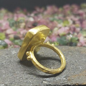 May include: A gold ring featuring a square-shaped gemstone. The ring is displayed on a dark grey surface with a backdrop of pink and green stones.