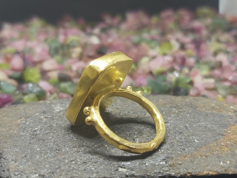 May include: A gold ring featuring a square-shaped gemstone. The ring is displayed on a dark grey surface with a backdrop of pink and green stones.