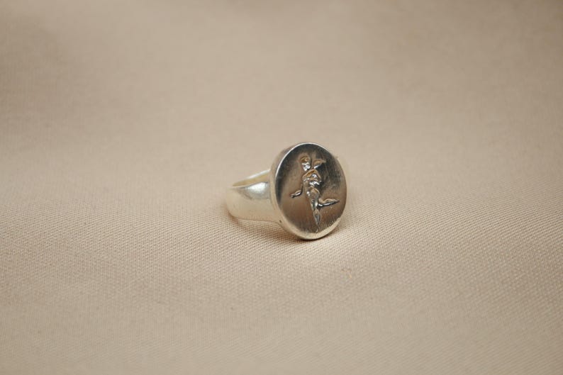 以下が含まれることがあります： 楕円形のトップにスタイリッシュな人物のレリーフデザインが施されたシルバーのシグネットリングです。