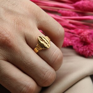Op de afbeelding: Een gouden ring met een getextureerd ovaalvormig bovendeel. De ring wordt aan een vinger gedragen.