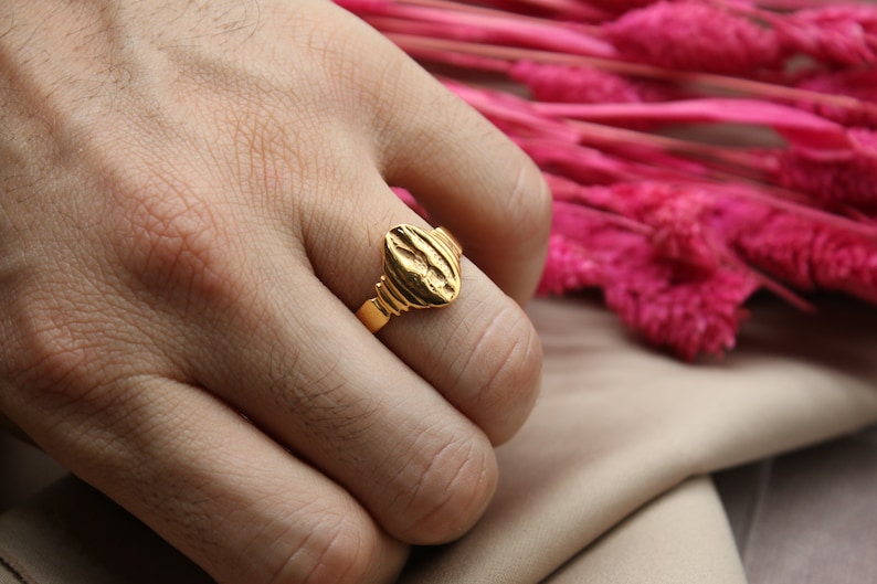 Op de afbeelding: Een gouden ring met een getextureerd ovaalvormig bovendeel. De ring wordt aan een vinger gedragen.