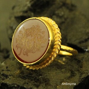 May include: A gold ring with an oval, light-colored stone featuring a detailed, golden design. The ring has a textured, ornate gold band and a reddish-brown border around the stone. The ring is set against a dark, textured background.