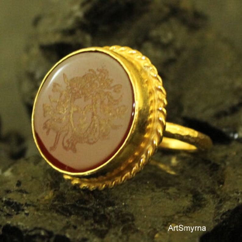 May include: A gold ring with an oval, light-colored stone featuring a detailed, golden design. The ring has a textured, ornate gold band and a reddish-brown border around the stone. The ring is set against a dark, textured background.