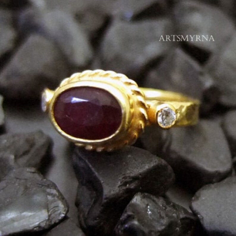 K&ouml;nnte beinhalten: Ein goldener Ring mit einem gro&szlig;en, ovalen Rubin und zwei kleineren, runden, wei&szlig;en Edelsteinen. Der Ring hat eine strukturierte Band.