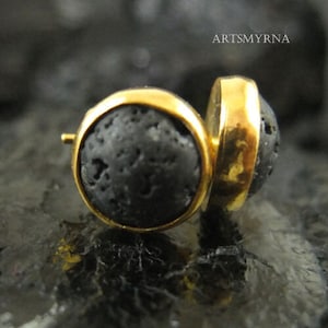 以下が含まれることがあります： 金色の枠にセットされた黒い溶岩の石のイヤリングのペア。イヤリングは丸く、ざらざらとした質感の表面をしています。