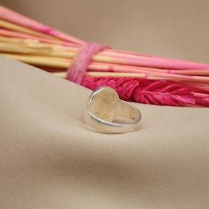 以下が含まれることがあります： 滑らかな楕円形のトップを持つシルバーリング。リングは薄い茶色の布地に置かれています。