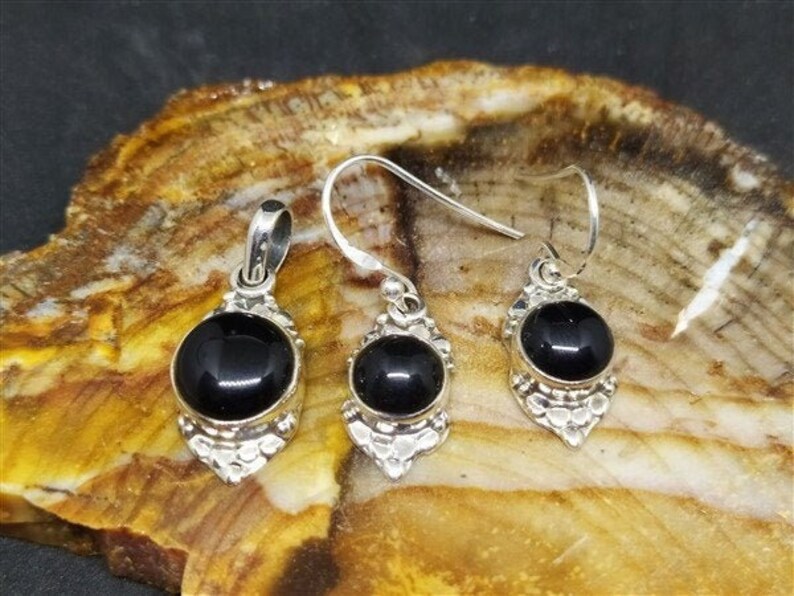 K&ouml;nnte beinhalten: Ein silbernes Anh&auml;nger- und Ohrring-Set mit schwarzen Onyx-Steinen. Die Steine sind in einem silbernen Filigran-Design gefasst. Der Anh&auml;nger und die Ohrringe befinden sich auf einem Hintergrund aus Naturstein.