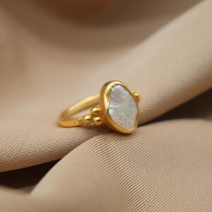 May include: A gold ring with a large, irregular-shaped white pearl. The ring has a textured band and small gold beads around the pearl.