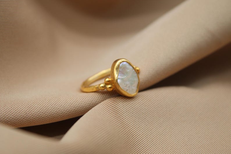 May include: A gold ring with a large, irregular-shaped white pearl. The ring has a textured band and small gold beads around the pearl.