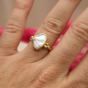 May include: A gold ring with a white, irregular shaped pearl-like stone. The ring is on a person's finger.
