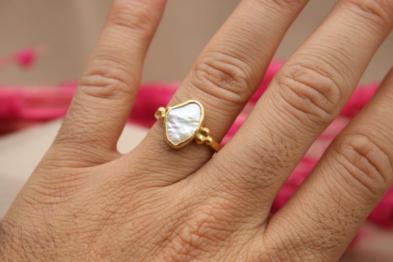 May include: A gold ring with a white, irregular shaped pearl-like stone. The ring is on a person's finger.