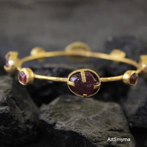 May include: Gold bangle bracelet with six red gemstone accents. The bracelet is made of hammered gold and has a simple, elegant design.